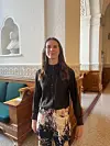 En kvinde står i en lys gang med buegange, grønne bænke og en buste i væggen.