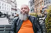 Skægget mand i læderjakke sidder på en bænk mellem cykler og bybygninger.