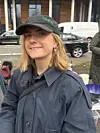 Kvinde i mørk jakke og kasket foran en bygning med biler i baggrunden.