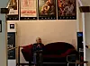Photo of an elderly woman sitting on a couch. Above her, movie posters line the walls. Taken inside Grand Teatret