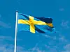 The Swedish flag waves from a white pole against a blue sky.