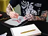 Tattooed hands sketch a red and black design on an iPad beside a notebook and pencil.