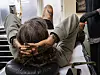 Gitte leans forward with her hands behind her head in a tattoo studio while Matilde works beside her.