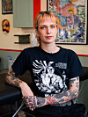 Matilde sits in a tattoo studio wearing a graphic T-shirt, with tattooed arms and artwork behind her.