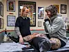 Gitte and Matilde discuss a detailed leg tattoo beside a tattoo table in a decorated studio.