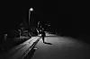 A man walks alone along a dimly lit, nearly empty street at night. He approaches a part of the street that is lit by a street lamp above him
