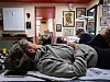 Gitte lays on a table in a room decorated with framed artwork, with another person seated nearby.
